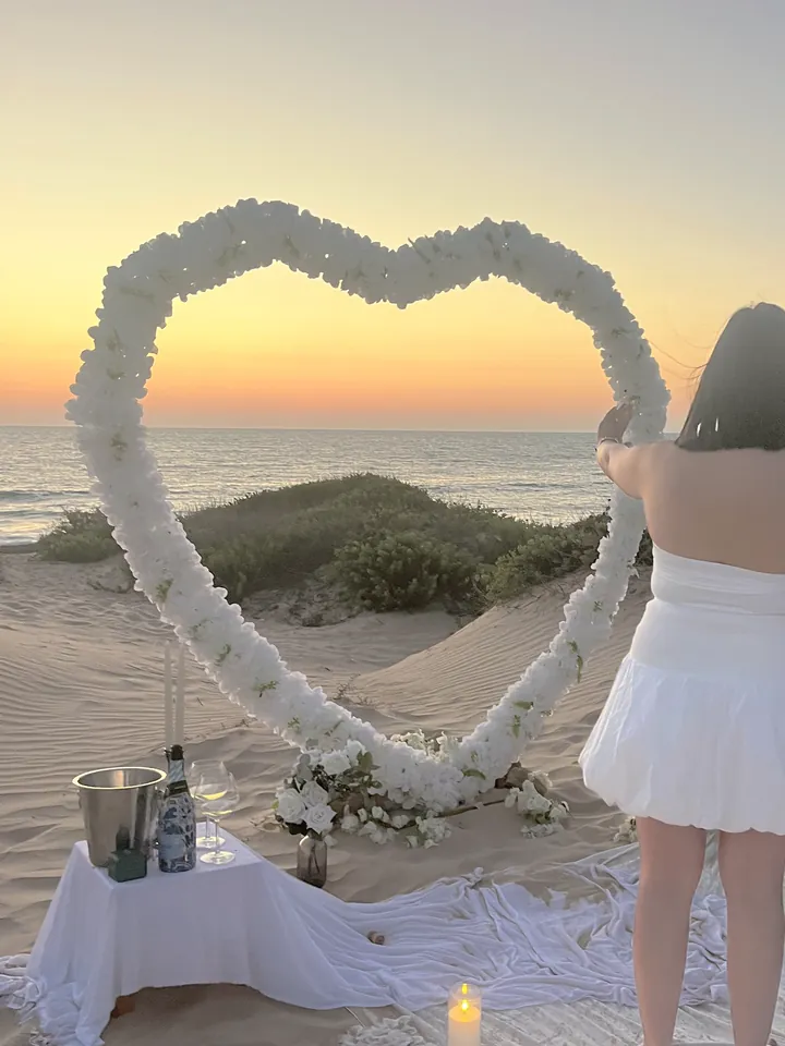 אישה עומדת מול קשת הלב מפרחים לבנים, שולחן שמפניה על החול